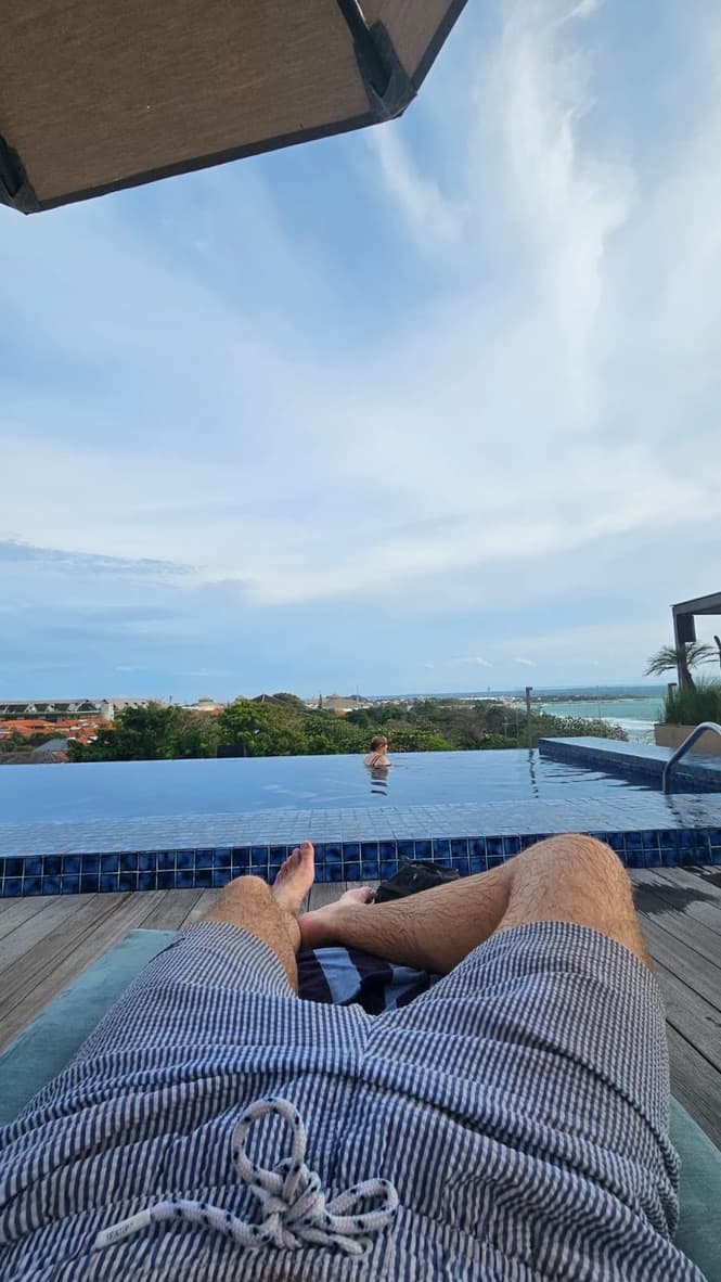 A student reclining on a lounger by an infinity pool in Bali, looking out over rooftops and greenery to the sea