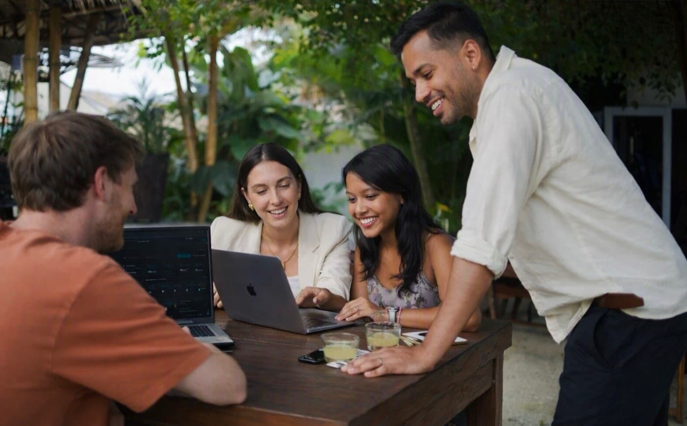 A supervisor leaning over a laptop with three interns smiling at an outdoor table in Bali